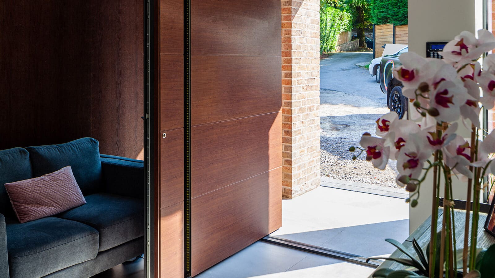 A modern home entryway featuring a large wooden pivot door opening to a sunlit driveway, with contemporary interior décor including a sofa, tiled flooring, and orchid arrangement
