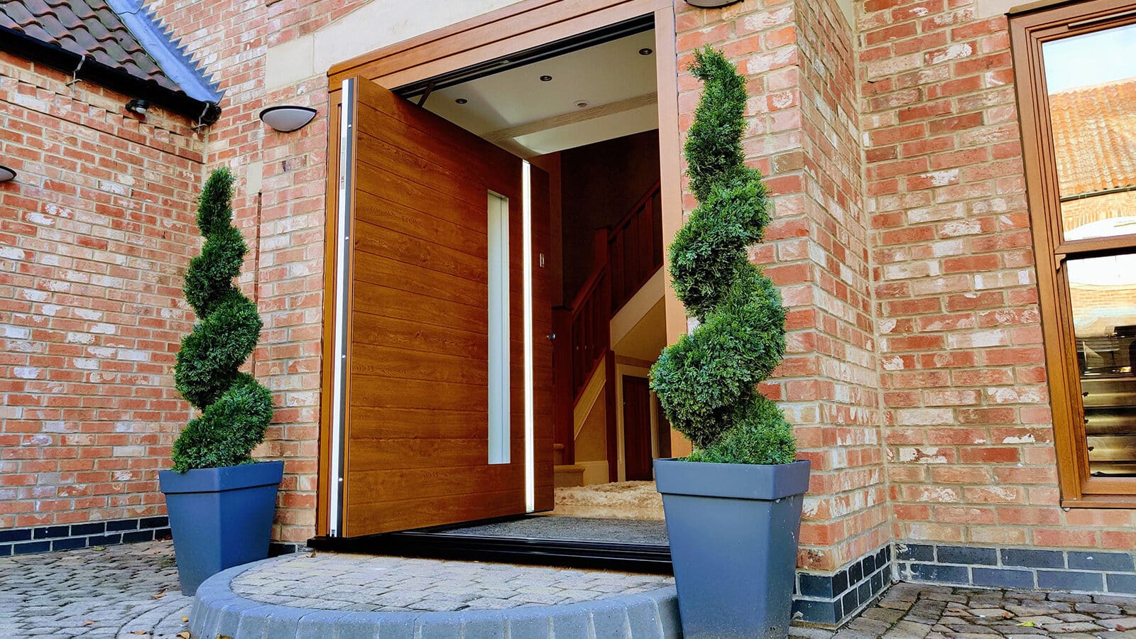 Wooden Spitfire S-700 pivot door set within a brick opening, shown with an exterior step and two plant pots